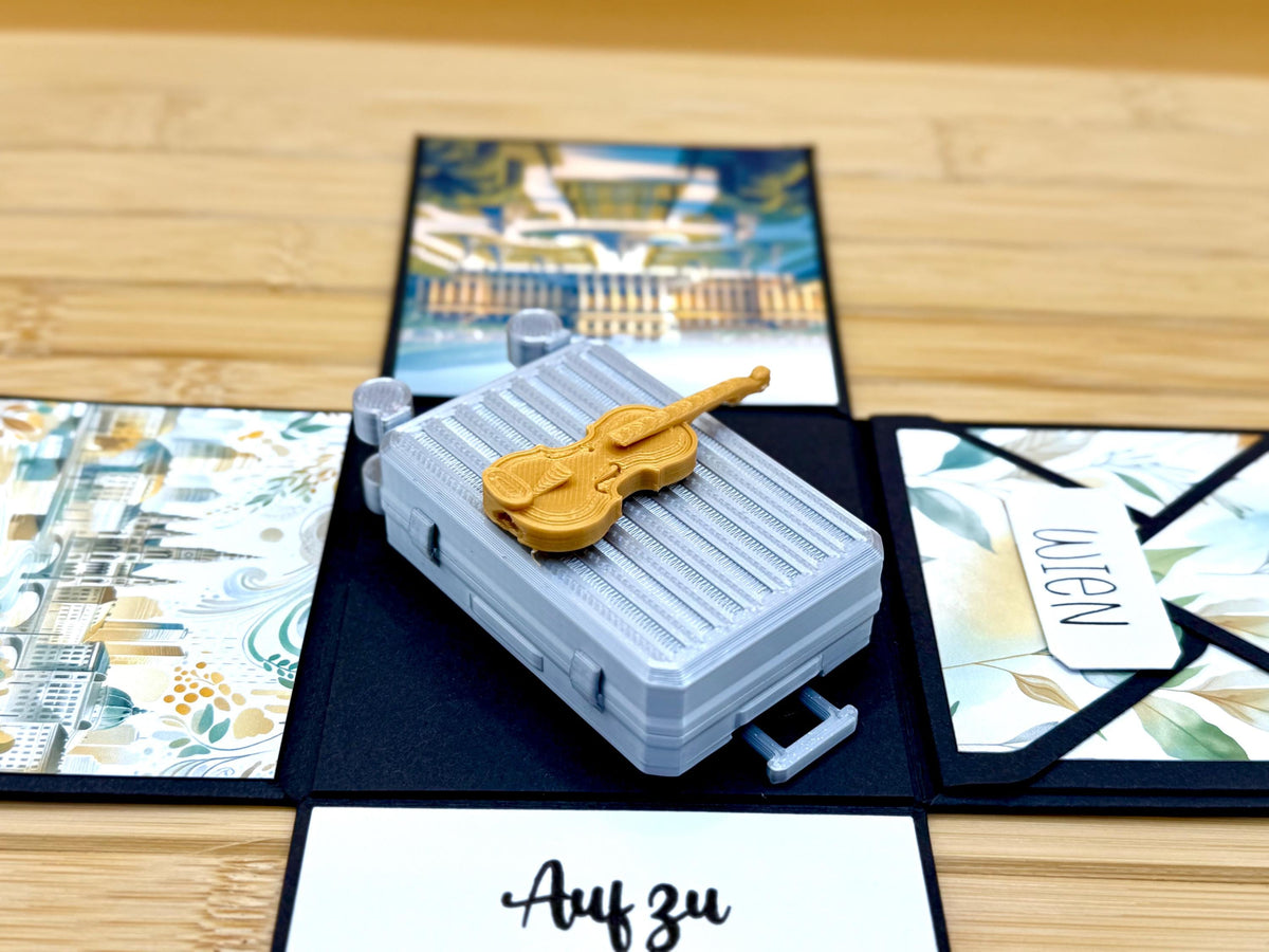 Nahaufnahme: 3D-gedruckter Miniaturkoffer mit goldfarbener Geige in der Explosionsbox Wien, rechts der Umschlag