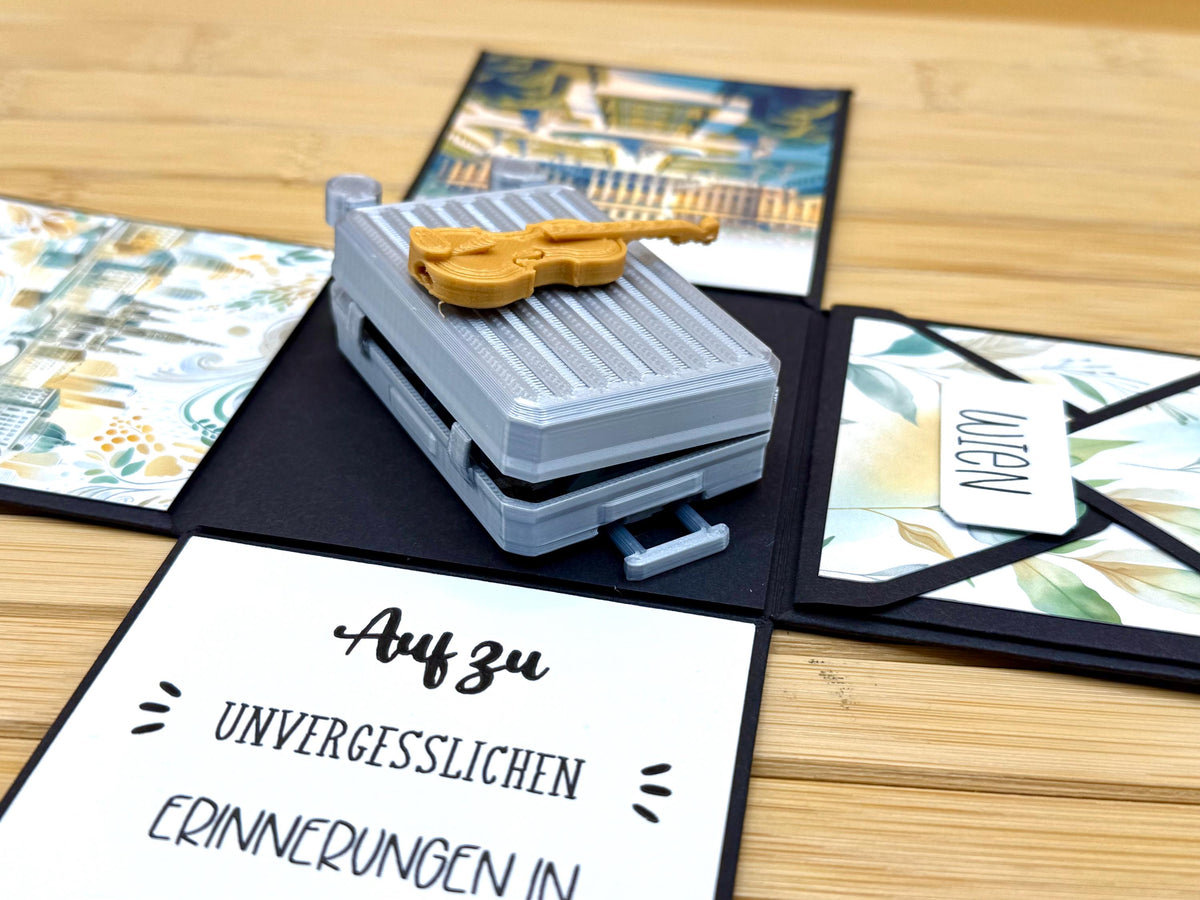 Explosionsbox Wien seitlich: Miniaturkoffer mit Teleskopgriff und Geige, vorn die Schriftkarte, rechts der Umschlag